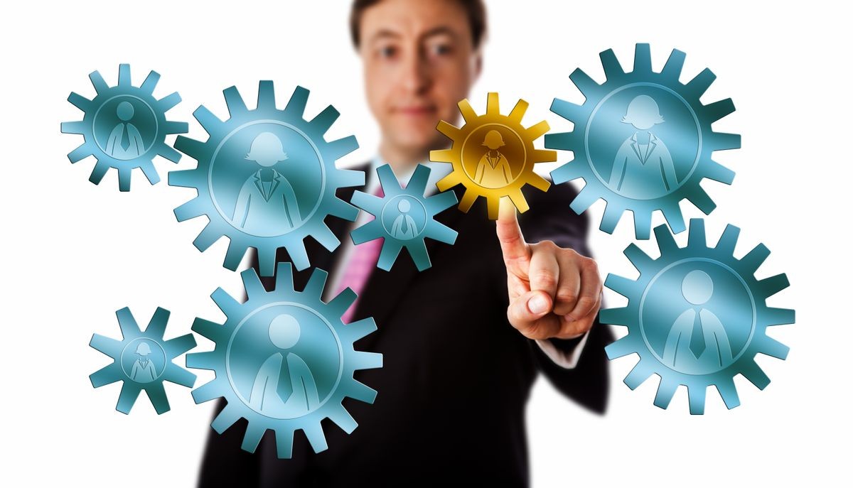 Smiling recruitment consultant is picking a female worker icon embossed on a golden cog in a mission critical position inside a virtual gear train of metallic cogwheels. Cutout isolated on white.